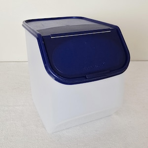 May include: A white plastic storage container with a dark blue lid. The lid has a curved design and a small, oval-shaped opening. The container is rectangular with rounded corners. The brand name "Tupperware" is visible on the lid.