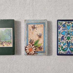 May include: Three decorative cards with different designs. The first card is green with a picture of a house and people in a park. The second card is blue and white with a floral design and bees. The third card is blue and white with a floral design and birds.