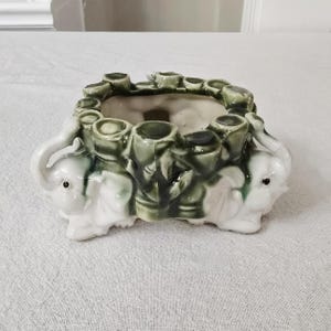May include: A rectangular ceramic planter with a white and green elephant design. The planter has a series of green cylindrical openings around the top edge. The elephants are white with gray details.
