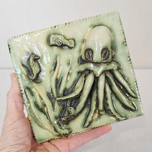 May include: Square ceramic tile with a raised relief design of an octopus, seahorse, and seaweed in shades of green and brown. The octopus has large eyes and tentacles. The tile is approximately 4 inches square.