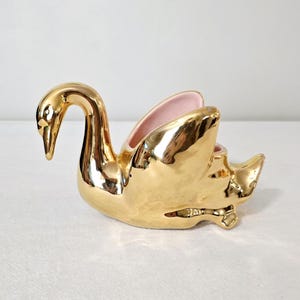 May include: A gold-colored swan-shaped ceramic planter with a glossy finish. The interior of the planter is a soft pink. The swan has a curved neck and detailed features, creating a decorative accent piece.