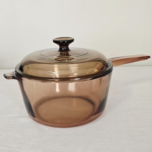 May include: A transparent amber-colored saucepan with a matching lid and handle. The saucepan has a rounded base and a handle extending from the side. The lid has a small, rounded knob. The cookware is made of glass.