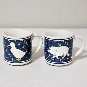 May include: Two white ceramic mugs featuring a dark blue background with a repeating white daisy pattern. One mug displays a white duck with a yellow beak and feet, while the other mug features a white pig. Both mugs have a white handle and rim.