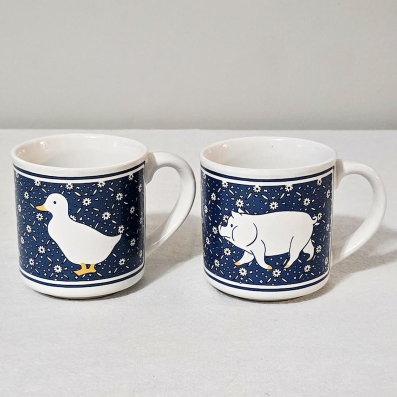 May include: Two white ceramic mugs featuring a dark blue background with a repeating white daisy pattern. One mug displays a white duck with a yellow beak and feet, while the other mug features a white pig. Both mugs have a white handle and rim.