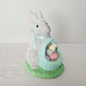 May include: A gray bunny figurine with a light blue ribbon around its neck, holding a light blue basket filled with pastel-colored Easter eggs. The bunny is sitting on a patch of green grass.