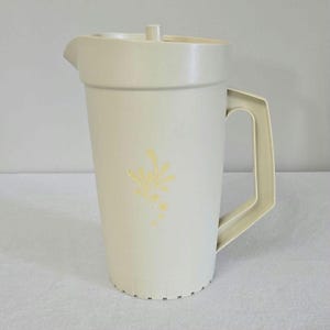 May include: Cream-colored plastic pitcher with a lid and handle. The pitcher has a floral design on the front. The pitcher is designed to hold liquids and has a spout for easy pouring.