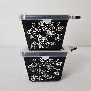 May include: Two black plastic containers with clear lids are stacked. Each container has a white floral design. The lids have a small handle on one side. The containers are square-shaped and appear to be food storage.