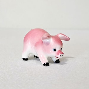 May include: A small ceramic pig figurine with a glossy finish. The pig is primarily light pink with darker pink shading on its back and ears. It features black painted hooves, a snout with two small nostrils, and dark eyes.