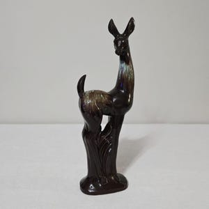 May include: A ceramic llama figurine in shades of brown, with a glossy finish. The llama has a long neck, upright ears, and a curved tail. The figurine is standing on a base with stylized grass.