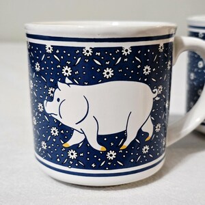 May include: A white ceramic mug featuring a dark blue background with a repeating pattern of small white flowers. A large, stylized white pig with yellow-tipped hooves is centered on the front. The mug has a sturdy handle and a smooth finish.