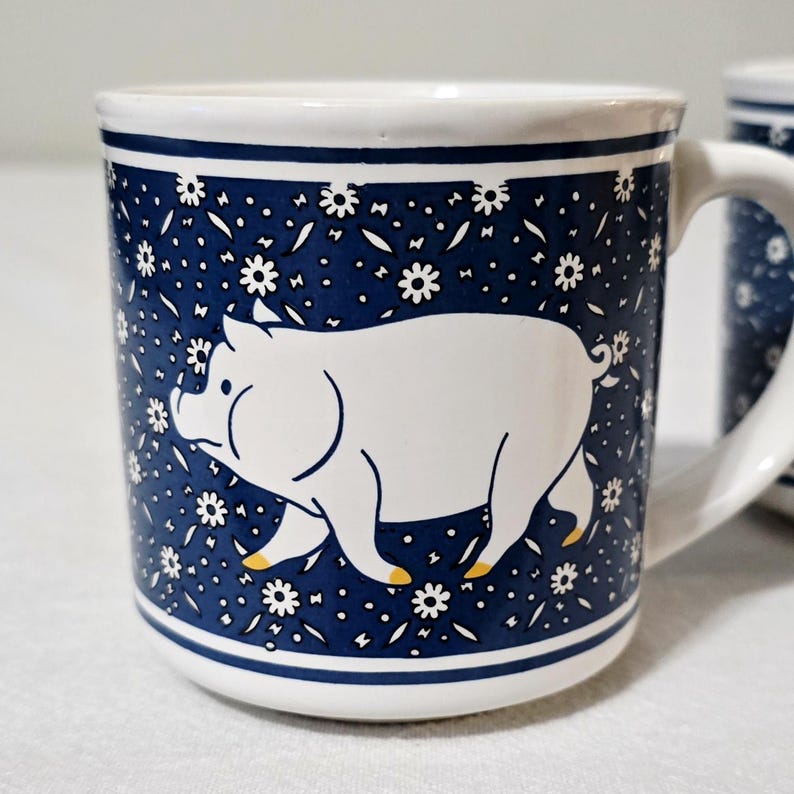 May include: A white ceramic mug featuring a dark blue background with a repeating pattern of small white flowers. A large, stylized white pig with yellow-tipped hooves is centered on the front. The mug has a sturdy handle and a smooth finish.
