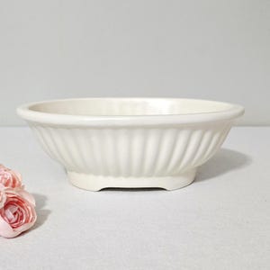 May include: A cream-coloured ceramic bowl featuring a wide, shallow basin and a distinct fluted or ribbed exterior design. The bowl has a smooth, glossy finish and sits on a slightly raised base, presenting a clean, uniform appearance throughout.