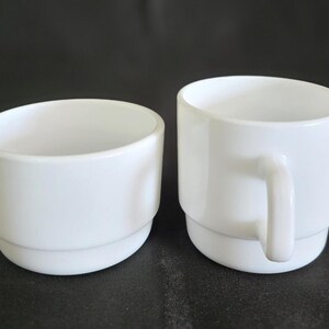 May include: Two white ceramic mugs, one with a handle and one without. Both mugs have a simple, rounded design.