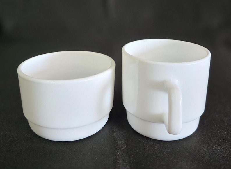 May include: Two white ceramic mugs, one with a handle and one without. Both mugs have a simple, rounded design.