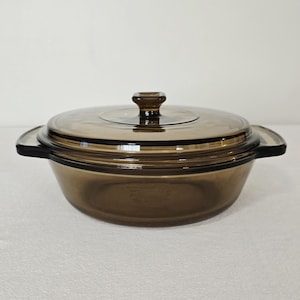 May include: A brown glass casserole dish with a matching lid. The dish has two handles and a knob on the lid. The glass is a translucent brown color. The dish is designed for cooking and serving.