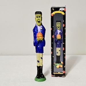 May include: A tall, slender figurine of a green-skinned character with dark hair, wearing a blue jacket and light-colored pants with black boots, holding an orange pumpkin. Beside it stands a black box labeled Hermitage Pottery Limited Edition.