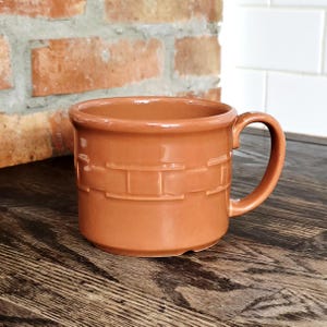 May include: A terracotta-colored ceramic mug featuring a smooth, glossy finish and a raised, embossed basket-weave pattern encircling the middle section. The mug has a wide, cylindrical body, a thick rim, and a curved handle on the right side.