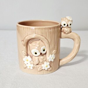 May include: A light brown ceramic mug with a textured tree bark pattern. The mug features a raised owl figure inside a hollowed-out tree section surrounded by white flowers. A smaller owl figure sits atop the handle, which is shaped like a branch.