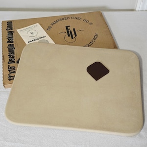 May include: A rectangular beige baking stone with rounded corners and a small dark brown scraper tool. The stone is positioned in front of its original cardboard packaging labeled The Pampered Chef Ltd. 12 inch by 15 inch Rectangle Baking Stone.