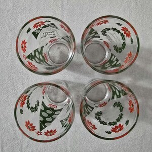 May include: Four clear glass tumblers with red and green Christmas-themed designs. Each glass features a red rim and is decorated with poinsettias, holly, and Christmas tree motifs. The glasses are arranged in a square.