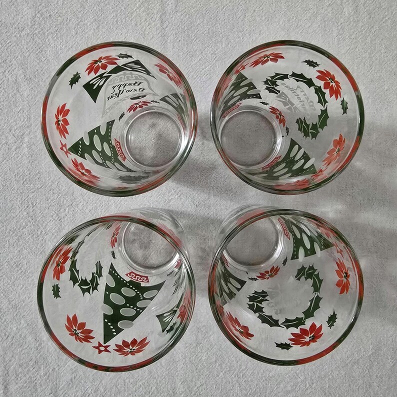 May include: Four clear glass tumblers with red and green Christmas-themed designs. Each glass features a red rim and is decorated with poinsettias, holly, and Christmas tree motifs. The glasses are arranged in a square.