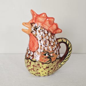 May include: A ceramic pitcher in the shape of a rooster. The pitcher features a red comb and wattle, with a body painted in shades of brown, yellow, and white. The handle is brown with gold and black accents. A decorative kitchenware item.
