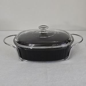 May include: Black oval casserole dish with a clear glass lid and silver-colored handles. The dish is sitting in a silver-colored metal stand. The casserole dish is on a light gray surface.