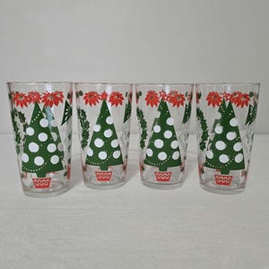 May include: Four clear drinking glasses with a Christmas tree design. Each glass features a green Christmas tree with white dots, red poinsettias, and green holly leaves. The base of each tree is in a red pot.