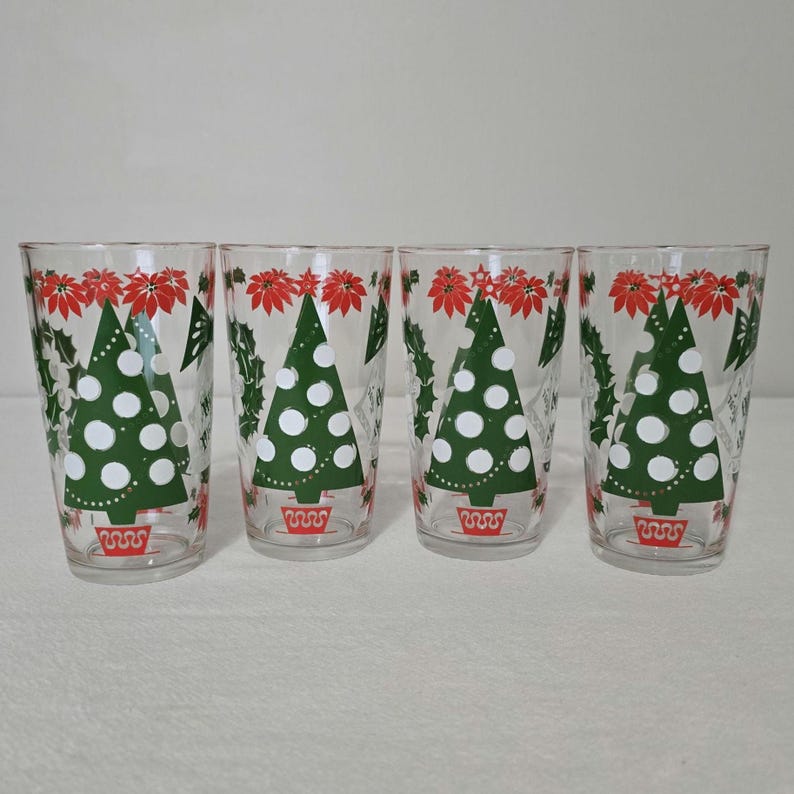 May include: Four clear drinking glasses with a Christmas tree design. Each glass features a green Christmas tree with white dots, red poinsettias, and green holly leaves. The base of each tree is in a red pot.