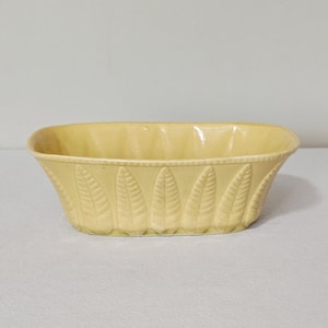 May include: A pale yellow oval ceramic bowl featuring a repeating embossed leaf pattern around the exterior sides. The rim has a subtle beaded texture, and the interior is smooth with a glossy finish, creating a consistent light yellow color throughout.
