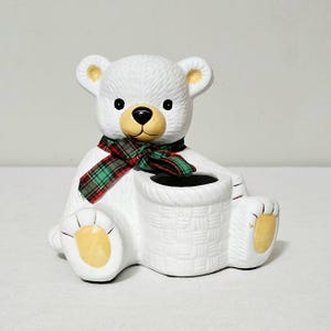 May include: A white ceramic bear figurine featuring a textured fur finish, a tan snout, and yellow paw pads. The bear wears a red and green plaid fabric scarf around its neck and sits beside a cylindrical basket with a woven pattern design.