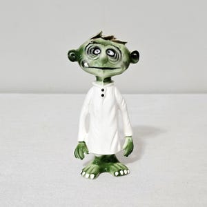 May include: A ceramic figurine of a green-skinned creature with a large head, wide eyes, and a prominent nose. The figure wears a long-sleeved white tunic with two small black buttons at the collar and has green hands and feet with white toes.