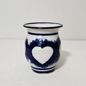 May include: A ceramic vase with a speckled off-white base and a dark blue painted band around the center. The blue band features a negative space heart shape in the middle. The vase has a flared rim and horizontal blue stripes at the top and base.