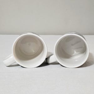 May include: Two white ceramic mugs positioned side by side, viewed from directly above. Each mug features a smooth, glossy white interior and a matching white handle extending to the side. The exterior rims show a hint of a dark gray pattern.