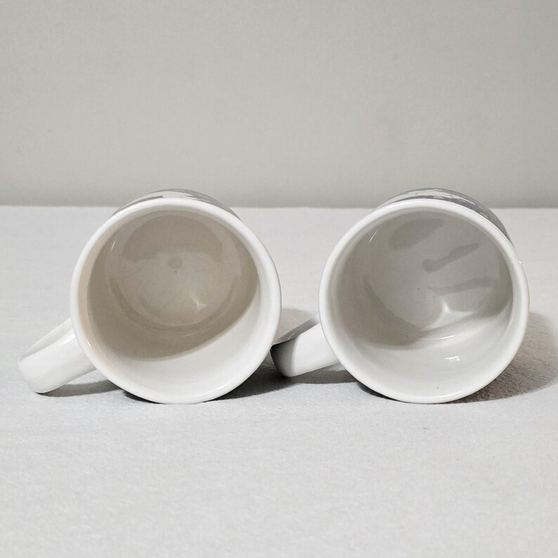 May include: Two white ceramic mugs positioned side by side, viewed from directly above. Each mug features a smooth, glossy white interior and a matching white handle extending to the side. The exterior rims show a hint of a dark gray pattern.