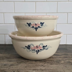 May include: Two cream-coloured ceramic mixing bowls stacked. Each bowl has a pink rim and a floral design in pink, blue, and grey. The bowls are on a wooden surface.