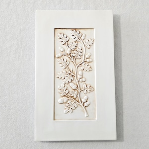 May include: A rectangular white ceramic wall tile featuring a central recessed panel with a raised relief design of an oak branch. The branch displays detailed oak leaves and several acorns, with subtle brown shading highlighting the relief texture.