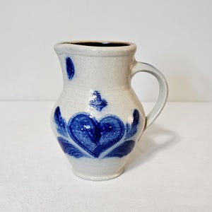May include: A speckled off-white ceramic jug featuring a blue hand-painted heart design on the front. The vessel has a curved handle, a wide spout, and a smooth glazed finish. The body is bulbous with a narrow neck and a flared rim.