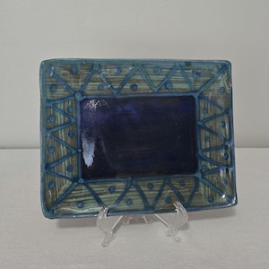 May include: A rectangular ceramic dish with a dark blue centre and a teal border. The border features a geometric design of triangles and dots. The dish is displayed on a clear plastic stand.
