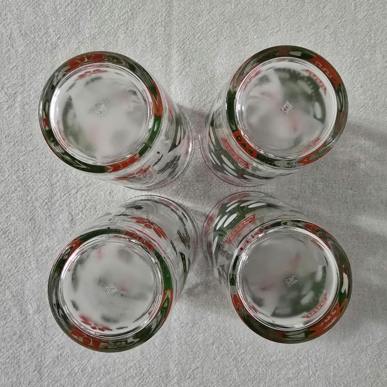 May include: Four clear drinking glasses with a red, white, and green design around the rim. The glasses are arranged in a square, with the bottoms facing the viewer. The design includes what appears to be a stylized floral pattern.