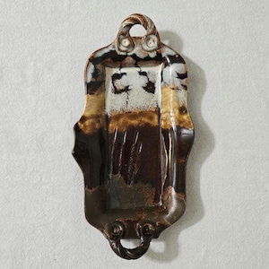 May include: A rectangular ceramic tray with a handle at each end and a loop at the top. The tray has a glossy finish with brown, white, and tan color variations. The design is organic and textured.