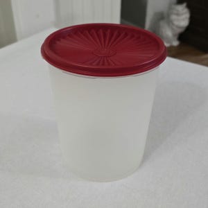 May include: A white, cylindrical container with a textured, red lid. The lid has a radial design emanating from the center. The container appears to be made of a translucent material, suitable for food storage.
