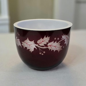 May include: A burgundy-colored bowl with a white interior rim. The bowl features a silver holly leaf and berry design around the exterior. The bowl is likely for decorative or serving purposes.