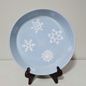 May include: A light blue ceramic plate with white snowflake designs. The plate is round with a slightly raised edge. The snowflakes are of varying sizes and shapes, adding a wintery theme.