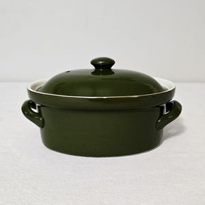 May include: An oval-shaped ceramic casserole dish with a glossy olive green finish. The dish features two integrated side handles and a matching removable lid with a central knob. The interior rim of the base is white, contrasting with the green.