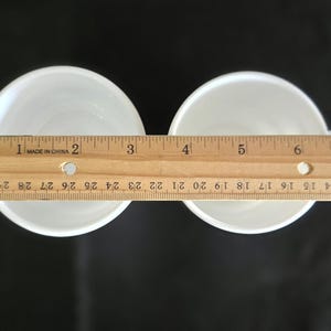 May include: Two white ceramic cups with a ruler measuring 6 inches across the top of the cups.