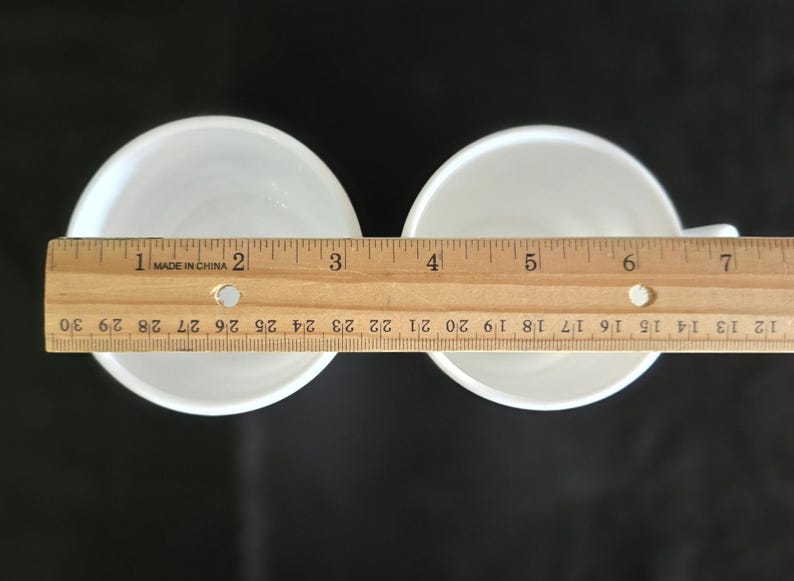 May include: Two white ceramic cups with a ruler measuring 6 inches across the top of the cups.