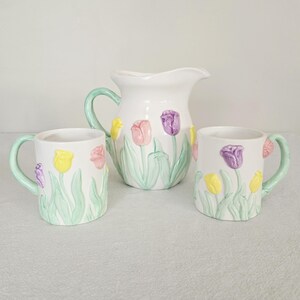 May include: A white ceramic pitcher and two mugs with a tulip design. The pitcher has a green handle and the mugs have green handles. The tulips are yellow, pink, and purple with green leaves. The set is likely for serving beverages.