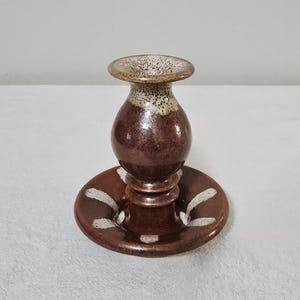 May include: A handcrafted ceramic candlestick holder. The holder features a dark reddish-brown body with a speckled tan top and white accents. The base is a round plate with white curved designs.