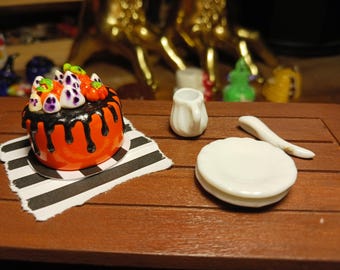 Könnte beinhalten: Ein Miniatur-Kuchen in Orange mit schwarzer Glasur, verziert mit kleinen Kürbissen und Geisterfiguren, auf einem schwarz-weiß gestreiften Papierquadrat. Daneben stehen ein kleines weißes Keramik-Kännchen, zwei gestapelte weiße Teller und ein weißer Löffel.